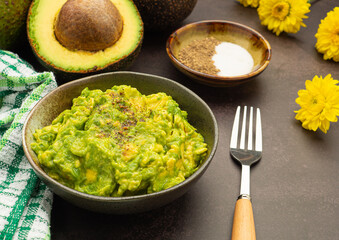 Fresh guacamole on a dish placed on a cutting wooden board with ingredients for homemade guacamole: avocados, lemon, salt, and pepper. Top view.  Concept of traditional Mexican preparation