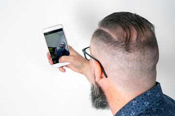 Bearded man using his smartphone to take a self-portrait, seen from behind and reflected on the cell phone