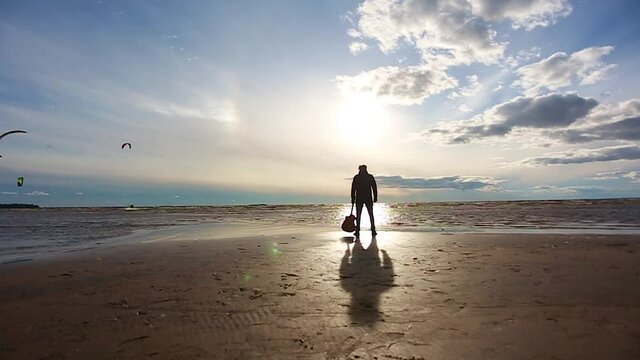 A man with a guitar stands with his back on the beach and looks at the waves of the ocean.