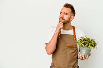 Young gardener tattooed caucasian man holding a plant isolated on white background looking sideways with doubtful and skeptical expression. - obrazy, fototapety, plakaty