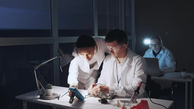 Mixed race electronics engineers in white coats working on motherboard using multimeter tester