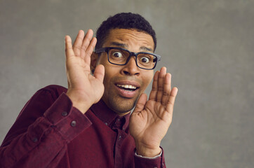 Studio portrait of black man in glasses gasping and panicking scared by something terrible. Shy African American guy afraid of camera looking at it with expression of fear and fright on his young face