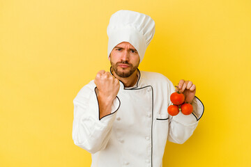 Young caucasian chef man holding tomatoes isolated on yellow background showing fist to camera, aggressive facial expression.