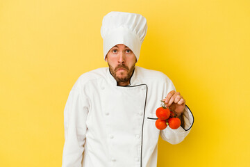 Young caucasian chef man holding tomatoes isolated on yellow background shrugs shoulders and open eyes confused.