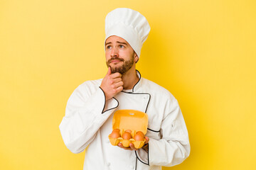 Young caucasian chef man holding eggs isolated on yellow background looking sideways with doubtful and skeptical expression.