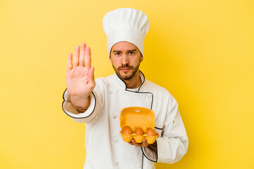 Young caucasian chef man holding eggs isolated on yellow background standing with outstretched hand showing stop sign, preventing you.