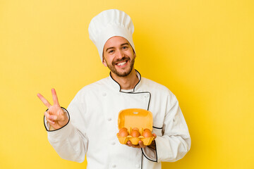 Young caucasian chef man holding eggs isolated on yellow background joyful and carefree showing a peace symbol with fingers.