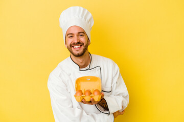 Young caucasian chef man holding eggs isolated on yellow background laughing and having fun.