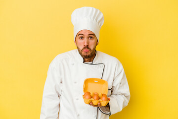 Young caucasian chef man holding eggs isolated on yellow background shrugs shoulders and open eyes confused.