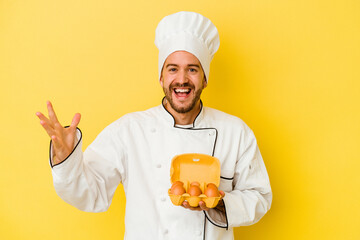 Young caucasian chef man holding eggs isolated on yellow background receiving a pleasant surprise, excited and raising hands.