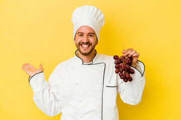 Young caucasian chef man holding grapes isolated on yellow background showing a copy space on a palm and holding another hand on waist.