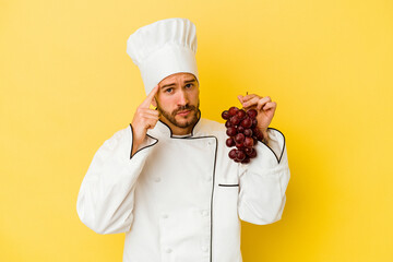 Young caucasian chef man holding grapes isolated on yellow background pointing temple with finger, thinking, focused on a task.