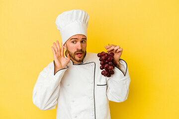 Young caucasian chef man holding grapes isolated on yellow background trying to listening a gossip.