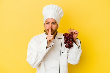 Young caucasian chef man holding grapes isolated on yellow background keeping a secret or asking for silence.