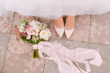 The bride's legs in graceful white shoes peek out from under the skirt, next to the bride's bouquet lies, close-up 