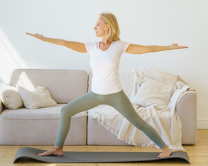 Calm and concentrated senior woman in home wear practicing Warrior Yoga Pose while exercising in...