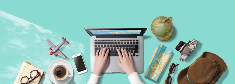 ノートパソコンをタイピングする手。海外旅行にまつわるアイテム。旅行の計画をたてているところ。もしくは、インターネットで旅行の疑似体験をしているところ。真上からのアングル