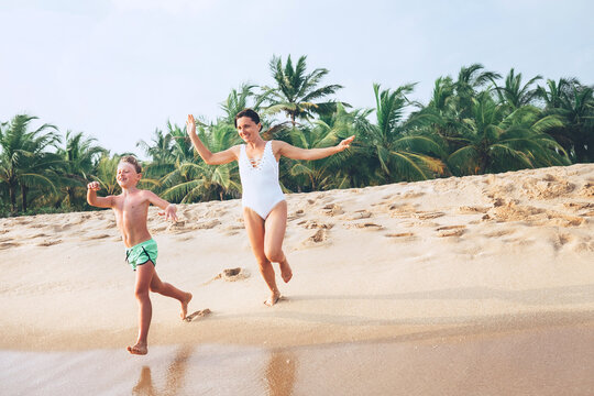 Mother and teenager son in swimsuits laughing and running by the perfect sand beach on tropical Sri Lanka island. Exotic countries traveling or sea vacation concept image.