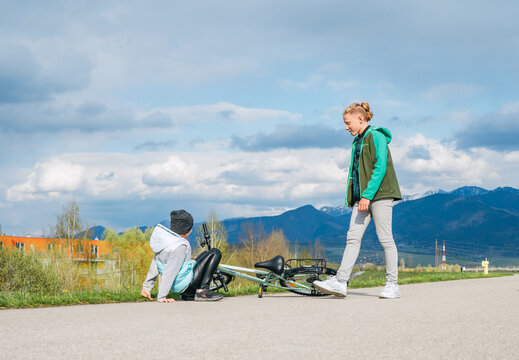 Brother helping his sister after bike falling while she doing first steps in riding. Happy childhood concept image. Kids on asphalt road with snowy mountains background.
