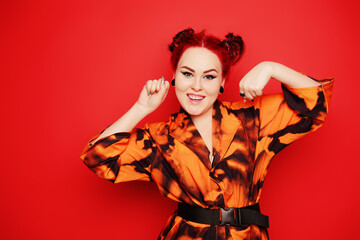 Cute girl on a red background. Red hair and tunnels in the ears, red dress and lips. Anime poses. The girl with eyeliner and two buns on her head grimaces, smiles, has fun. Japanese culture