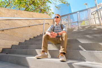 A young blond man in a shirt rests sitting on the steps of a building on a sunny summer day