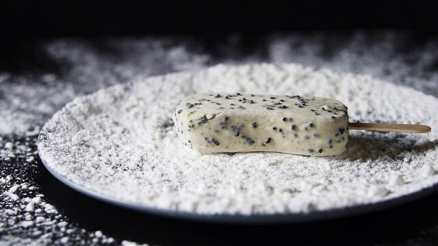 White ice cream sundae falls into white powder in a plate. Powdered sugar is scattered over a black background. Falling chocolate into a plate