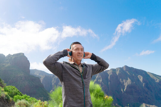 Happy mature man traveler in headphones standing on top of hill and enjoying beautiful mountain landscape