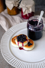 Cheesecakes cottage cheese fritters with berry jam topping, jar of jam with a spoon inside. Russian sweet breakfast cheese pancakes
