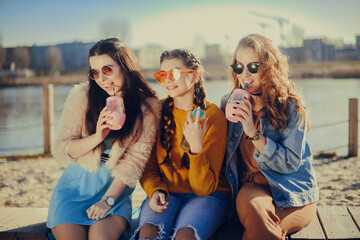 Three girls sit on the beach , near the river, talk, gossip, joke, laugh, smile at womans day. Girls on a hot day eat ice cream. Beauty model wearing sunglasses at summer day. Girl with hairslyle - obrazy, fototapety, plakaty