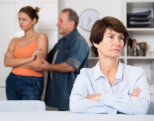 Upset mother is sitting at the table and her family is sympathying with her at home.