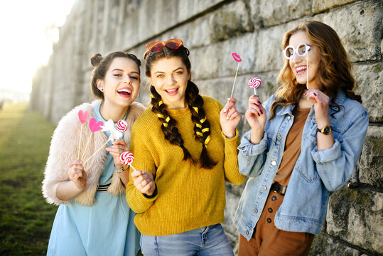 Hipster girls, colored dressed, with sunglasses and wrist watch. Grup of three girls with funny lips, glasses, stars, paper hearts on stick at the sunset having fun. Going crazy, smile at summer day.