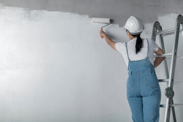 girl paints a white wall with a roller. Repair of the interior. Young female decorator painting a wall in the empty room, concept builder or painter in helmet with paint roller over the empty room