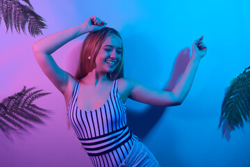 Obraz na płótnie Canvas A model in neon poses. Fashion photo of a girl in a swimsuit and shorts in pink-blue light. Girl on a background of tropical leaves summer photo. Beautiful blonde