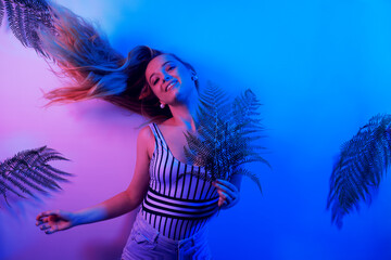 Dance party summer vibe. A model in neon poses. Fashion photo of a girl in a swimsuit and shorts in pink-blue light. Girl on a background of tropical leaves summer photo.