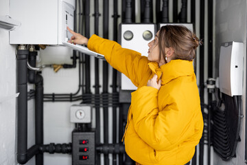 Young woman feeling cold, wearing jacket in the boiler room. Housewife adjusts temperature on the gas boiler at home