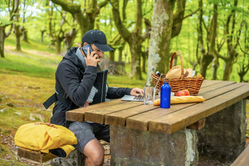 A young Caucasian father teleworking seated at a picnic table with the computer, with the child in the backpack. Summer lifestyle