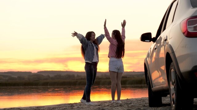Free women travelers stand next to car on beach enjoying sunset in park, jumping and clapping rejoicing. Girls drivers stopped at campsite. Girlfriends are enjoying trip in car. Vacation, adventure