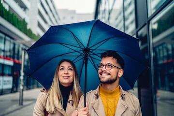 Happy young couple standing under an umbrella and looking around the city.