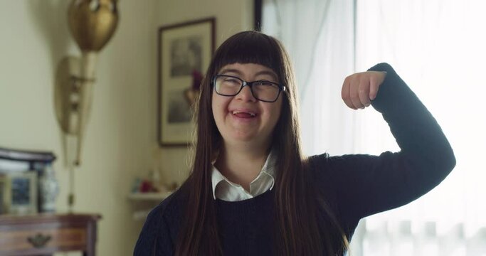Authentic shot of happy friendly teen girl with down syndrome is showing her power with arms and smiling in camera at home. Concept of handicapped, child care, healthcare, disability, people, optimism