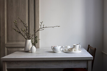 Half-open branches of an apple tree in a white fluted vase. A small vase with a branch of dried eucalyptus and two cups of coffee on a tray on the table.