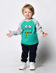 small smiling preschooler of European descent in a trendy green raglan with cute black sweatpants with a print on the face stands and spreads his arms. Studio portrait on white