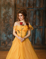 Girl beauty fantasy princess in yellow long historical, medieval silk dress holding flower red rose in her hands. Background of old gothic castle room. Fairy tale bewitched queen. Happy woman smiles © kharchenkoirina