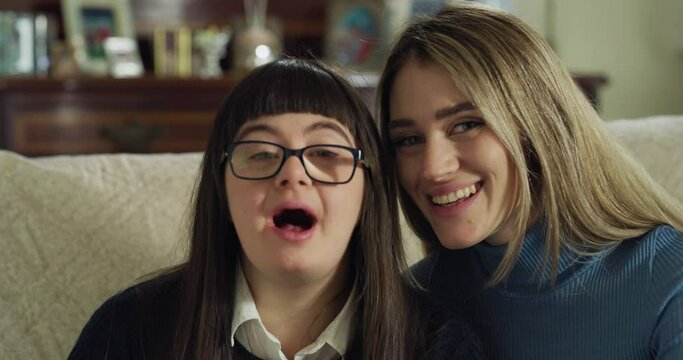 Authentic shot of happy young woman embracing her teen sister with down syndrome and smiling in camera at home. Concept of love, family, handicapped, healthcare, disability, love, friendship