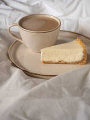 Morning cup of coffee and classic cheesecake on a beige ceramic plate on a soft white bed in the morning light