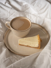 Morning cup of coffee and classic cheesecake on a beige ceramic plate on a soft white bed in the morning light