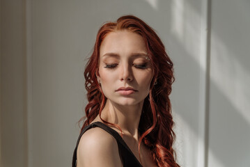 Fototapeta premium an attractive red-haired woman with her eyes closed enjoys the sunlight from the window. close-up portrait of a beautiful woman with her eyes closed. the concept of calm, harmony
