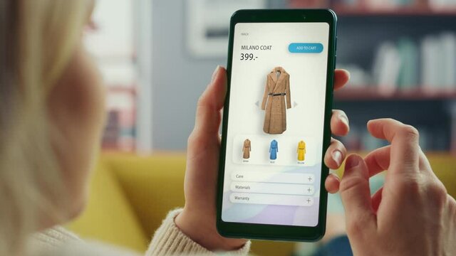 Beautiful Authentic Female Online Shopping on Her Smartphone at Home Living Room while Lying on a Couch Sofa. She's Browsing the Internet and Checking Deals in Her Favourite Internet Clothing Store.