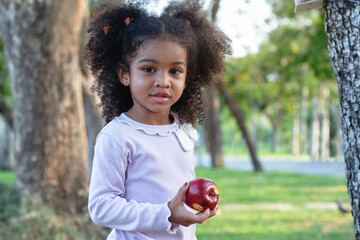 Cute curly hair girl play in green park, 3 years old, holding red apple and looking at camera