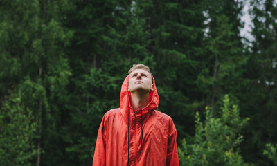 Portrait of a young male tourist in a red raincoat stands in the rain in the mountains against a...