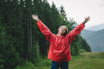 Joyful tourist girl standing in mountains in rainy weather against beautiful landscape background, looking into camera and smiling with arms raised. Happy female tourist enjoys the rain on a hike.